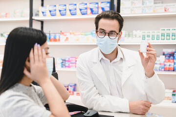 Obraz premium Male pharmacist druggist in face mask against coronavirus advising remedy, painkillers, drugs, medicines, pills for female customer client buyer suffering from headache migraine in pharmacy drugstore