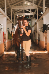 Young woman rider in equestrian uniform standing in stable next to her horse and petting it. Horseback riding training. Love and affection for domestic brown horse. Authentic village barn.
