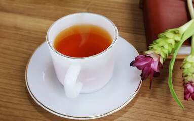 Hot tea in a pearl cup with a saucer on the side. A book with a long bouquet of red and green flowers on a wooden table.

