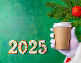 Coffee shop, cafe concept. New Year postcard. 2025 numbers written as ginger bread. Santa Claus holding takeaway paper coffee cup in a hand, wearing white gloves, on green background, closeup.