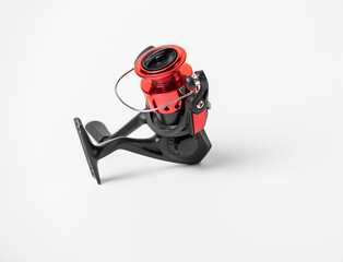 A red and black fishing reel sits on a white background. The reel is small and has a black handle