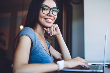 Portrait of happy skilled female student enjoying e learning in coworking space, successful hipster girl sitting at desk with laptop device and looking at camera during break from distance job