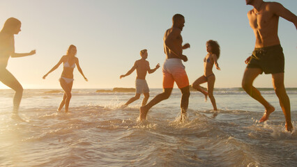 Group Of Friends In Swimwear Run Through Waves At Sunset On Summer Beach Vacation With Lens Flare