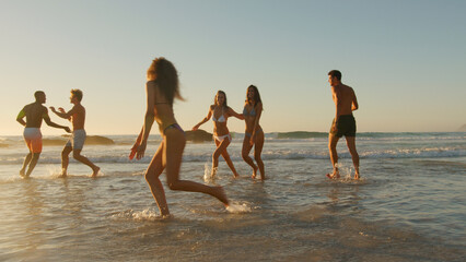 Group Of Friends In Swimwear Run Through Waves At Sunset On Summer Beach Vacation With Lens Flare