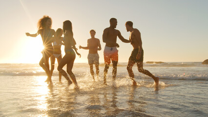 Group Of Friends In Swimwear Run Through Waves At Sunset On Summer Beach Vacation With Lens Flare