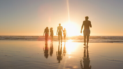 Fototapeta premium Group Of Friends In Swimwear Walk Towards Waves At Sunset On Summer Beach Vacation With Lens Flare