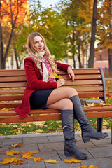 Fototapeta premium A beautiful woman in a red coat sits on a bench in an autumn park. She is wearing black boots and a scarf.