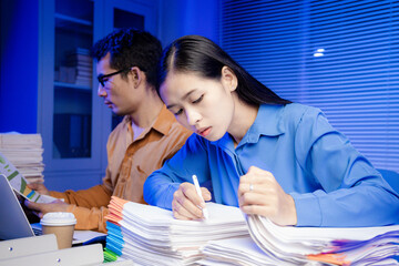 Two Asian women work late into the night. They are exhausted and stressed at their desks. They are...