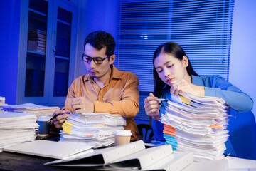 Two Asian women work late into the night. They are exhausted and stressed at their desks. They are...