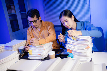 Two Asian women work late into the night. They are exhausted and stressed at their desks. They are...