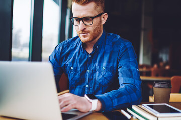 Concentrated male freelancer searching information for creating and programming new website for company order, serious hipster guy in optical eyeglasses reading news about internet provider via laptop