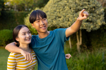 Fototapeta na wymiar Smiling young asian brother and sister embracing and pointing at something outdoors in park