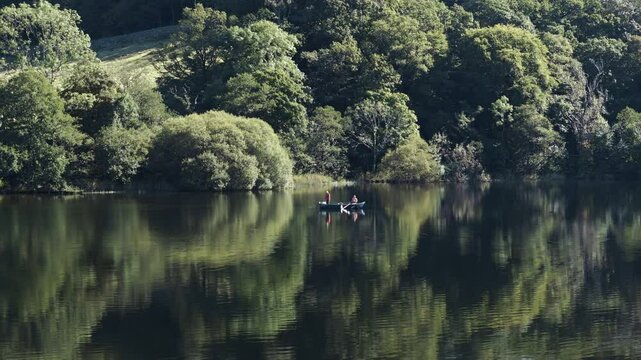 Aerial orbit of fishers on Grassmere Lake in Lake District, England, at a scenic spot with serene lush forest vegetation reflecting in water as they row