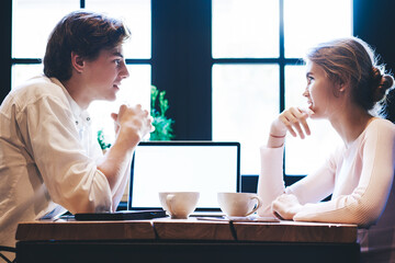 Young caucasian man and woman sitting at cafeteria table with laptop computer with mock up screen for text, millennial female students socialising in coffee shop discussing modern technology