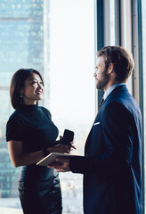 Successful diverse male and female employees in formal wear discussing financial ideas while standing near office window of skyscraper building. Corporate man and woman colleagues talking about work