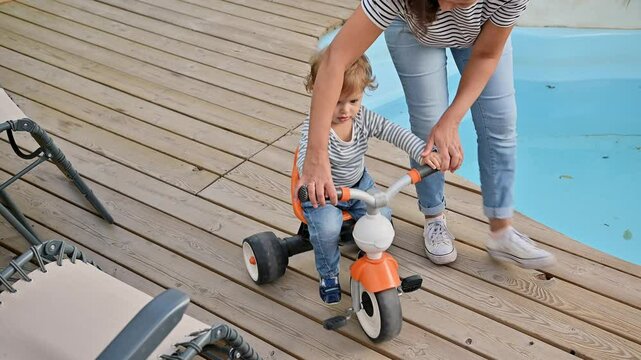 Child learning to ride a tricycle with parent by the poolside, wooden deck creates a warm and inviting outdoor scene, perfect for family bonding and outdoor activities