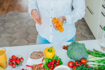 Female hands holding pill organizer with nutritional supplements on table with fresh food...
