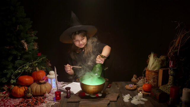 Halloween, smiling little cute girl dressed as witch in witch's hat brews a glowing magic potion in cauldron on coals. Magic wand, сandles, pumpkins, ingridients, fog smoke, magic, mystical atmosphere
