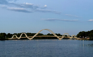 Obraz premium wavy bridge structure spanning across a calm body of water. The bridge’s smooth, flowing arches contrast with the serene surroundings of trees and a clear blue sky, creating a striking and modern desi