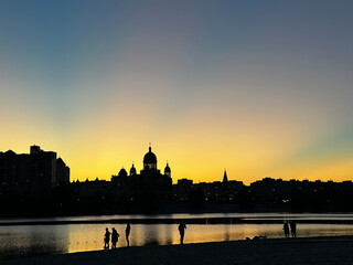 Obraz premium peaceful sunset by a river, with silhouettes of buildings, including a church with domes, against a golden sky. People stand by the water’s edge, enjoying the serene evening scene.