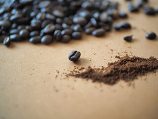 Auf einem hellbraunen Untergrund liegt ein Haufen Kaffeebohnen, eine einzelne Bohne liegt davor, daneben gemahlenes Kaffeepulver
