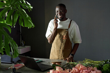 Waist up portrait of smiling African American man speaking on phone and looking at laptop screen while managing flower shop with elegant dark setting