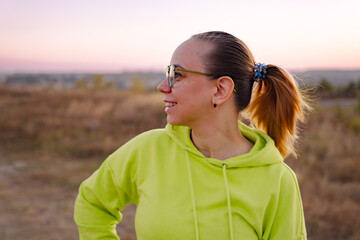 A woman with glasses stands outdoors, gazing thoughtfully toward the horizon as the sun sets, casting warm colors across the tranquil landscape.