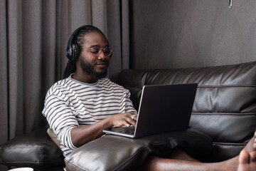 Young African Man Working Online from Home on Laptop, Relaxed on Couch with Headphones, Modern Home Office Setup