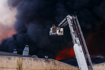 Obraz premium Firefighters use telescopic tower during massive large blaze fire, firetruck with crane, firemen team putting out the fire with extendable fire ladder, blazing warehouse factory, burning building