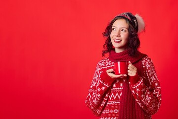 A young woman in a red sweater with snowflakes and a red hat with a white pom-pom, smiling and looking to the side. She holds a mug with a red handle. The background is solid red.