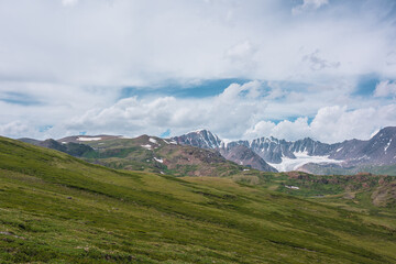 Obraz premium Scenic alpine landscape with green hills and rocks with view to large snow-capped mountain top and big glacier far away under clouds in blue sky in changeable weather. Awesome high snowy mountains.