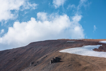 Rock and snowfield on mountainside in changeable weather. Sunlit stony mountain range with snow under clouds in blue sky. Sunlight and shadows on stone hill in sunny day. Rocky hillside in bright sun.