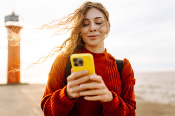 Young woman enjoying nature by the waterfront during a cloudy day by a lighthouse. Phone call by the sea. Travel, tourism, technology, blogging concept.