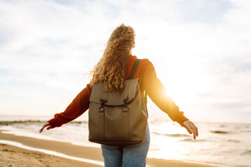 As she walked along the beach, the woman tourist adjusted her backpack, taking in the salty sea air and the sound of the waves at sunset.  Calmness and tranquility. Travel, tourism concept.