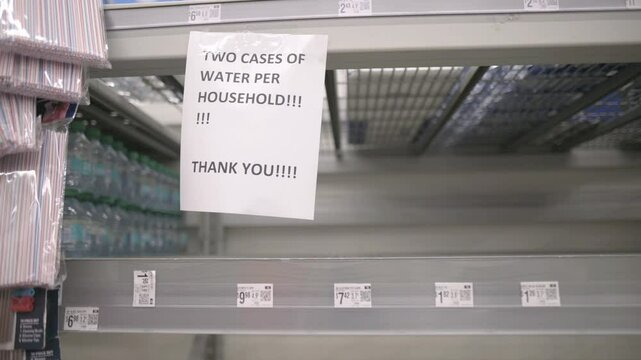 Empty water shelves in a supermarket, with a rationing sign limiting two cases per household after a hurricane. Reflects supply shortages and emergency measures. High quality 4k footage