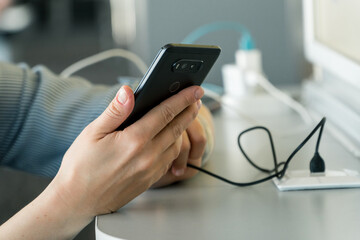 Close up of female hands charging smartphone using cord and usb plug in a public space while using a phone.