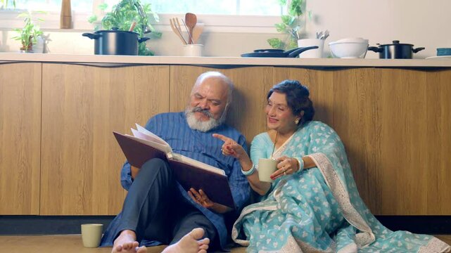 Joyful Indian Asian senior couple sitting comfortably on the kitchen floor, relaxing with cups of tea,fondly looking through an old photo album, reliving cherished memories together