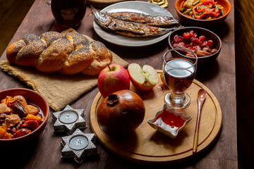 Challah, honey, apple and pomegranate and wine and chelnt and fish on the table with menorah and burning candles in honor of Rosh Hashanah