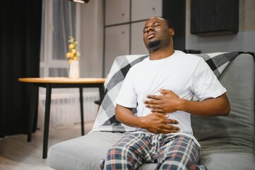 Unhappy guy of african american ethnicity, sits on a sofa in the living room, holds his hands on his stomach, grimaces from pain in his stomach