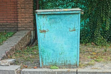 one retro green iron rusty big lidded heavy industrial square urban painted old dirty trash can stands on road near brick fence daytime outdoors