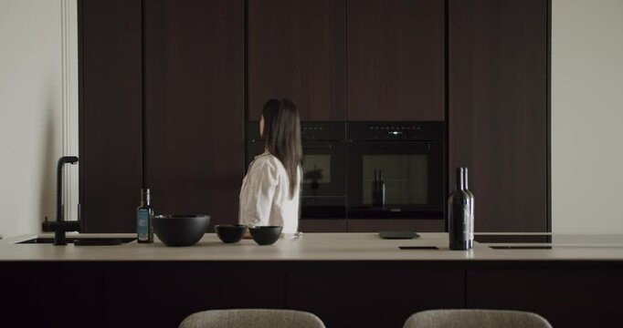 Woman standing in a modern kitchen with dark cabinetry, minimalist decor, and kitchenware on the counter. Woman cooking in a modern kitchen with a dark interior design, bottle of wine on counter.