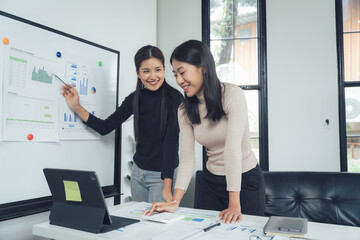 Collaborative Success: Two smiling Asian businesswomen review data on a whiteboard and laptop, showcasing teamwork and strategic planning in a modern office. 