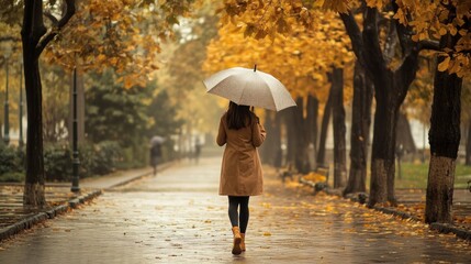 Obraz premium Woman walking alone in a park with an umbrella during a fall day.
