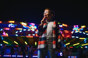 A cute caucasian boy eating a colorful lollipop standing against the background of a carousel in...