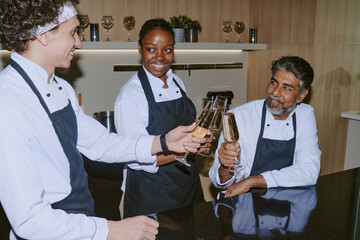 Happy cooking team in white uniform with black aprons holding glasses with champagne and clinking them