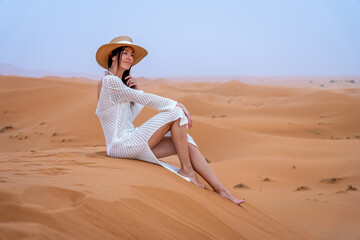 Young Brunette Woman in Camel Hat and White Dress Sitting on Erg Chebbi Dunes at Sunset