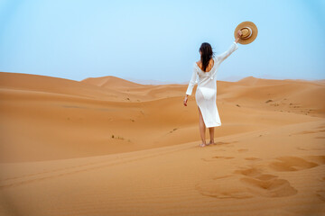 Young Brunette Woman in Camel Hat and White Dress Waving from Behind in Erg Chebbi Dunes