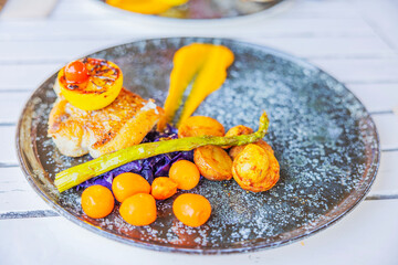 Gourmet fish dish served on plate with roasted potatoes, asparagus, cherry tomatoes, purple cabbage, and grilled lemon, presented on white table. Curacao.