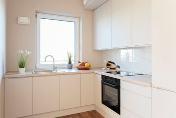 Modern kitchen interior with kitchen sink near window with faucet, beige cabinets and stylish furniture with oven and hob. New apartment and real estate.