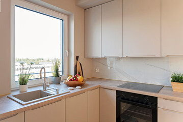 Modern kitchen interior with kitchen sink near window with faucet, beige cabinets and stylish furniture with oven and hob. New apartment and real estate.
